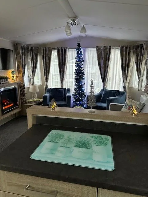 Lounge at Tern 32 with a slim blue-lit Christmas tree between two blue armchairs, warm lamps, light-up trees on the sideboard, and the kitchen worktop in the foreground