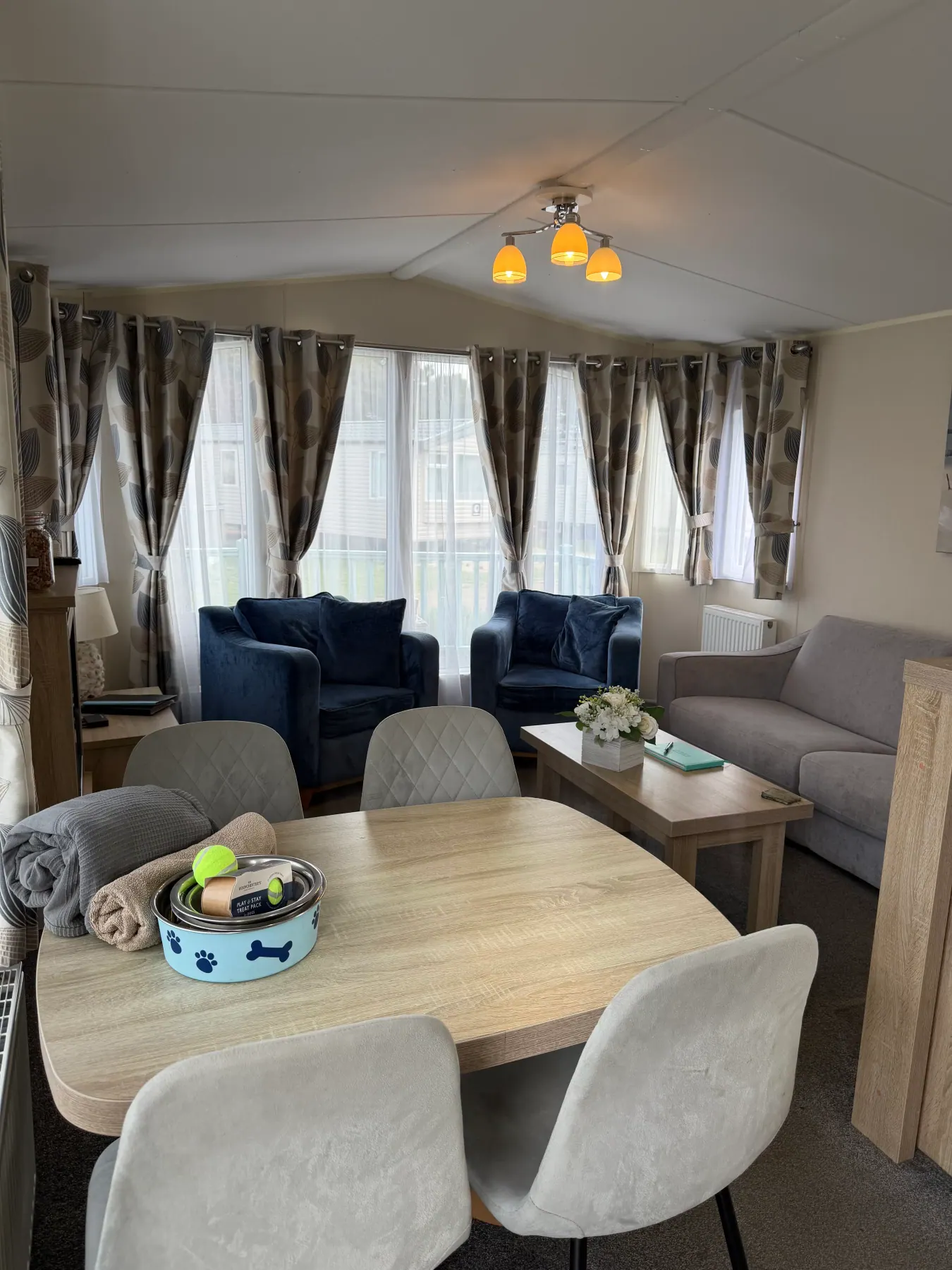 Open-plan living and dining area inside Tern 32 with a wooden table for six, a grey sofa, two deep blue armchairs and dog bowls