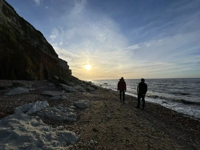 Two people walking below the cliffs on a beach at sunset, with the low sun over the sea