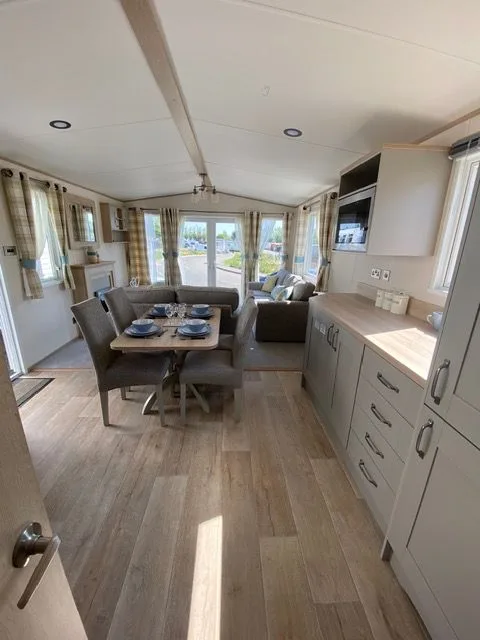 The open-plan living and kitchen area inside Windsor 30, seen from the doorway, with a dining table laid for four, grey seating, pale grey units and wood-effect flooring